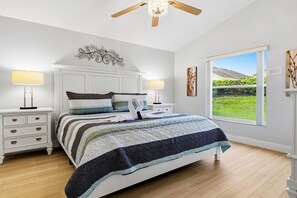 A bedroom with a bed, two bedside tables, a ceiling fan, and a window with a view of greenery.
