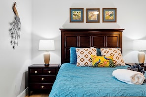 A bedroom with a wooden headboard, a blue bedspread, patterned pillows, a white throw, and a nightstand with a lamp.