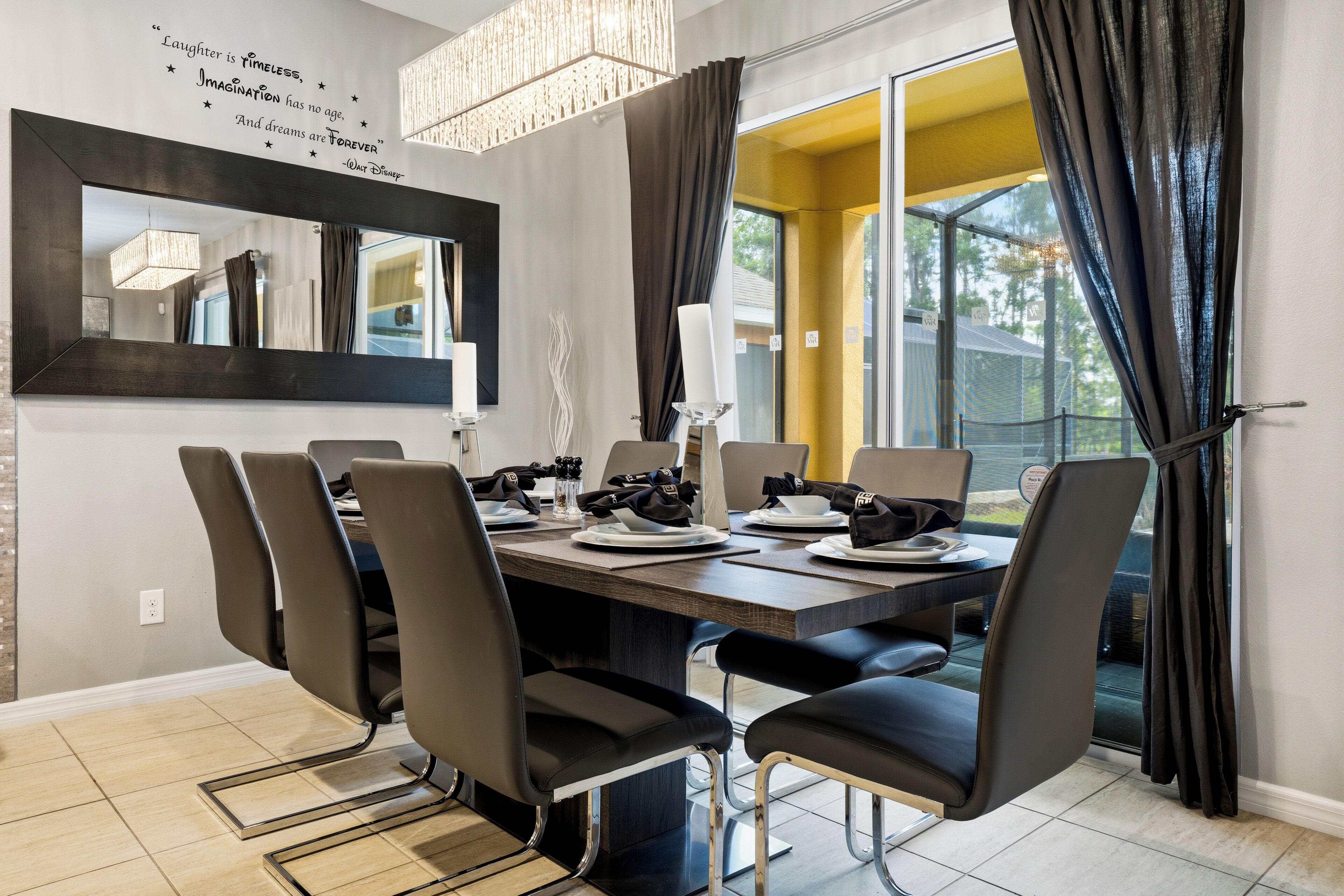 A dining area with a large table, chairs, a chandelier, and a mirror on the wall.