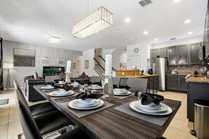 Una cocina moderna con electrodomésticos de acero inoxidable, un comedor con una larga mesa dispuesta para seis personas, y una escalera que conduce a un nivel superior.