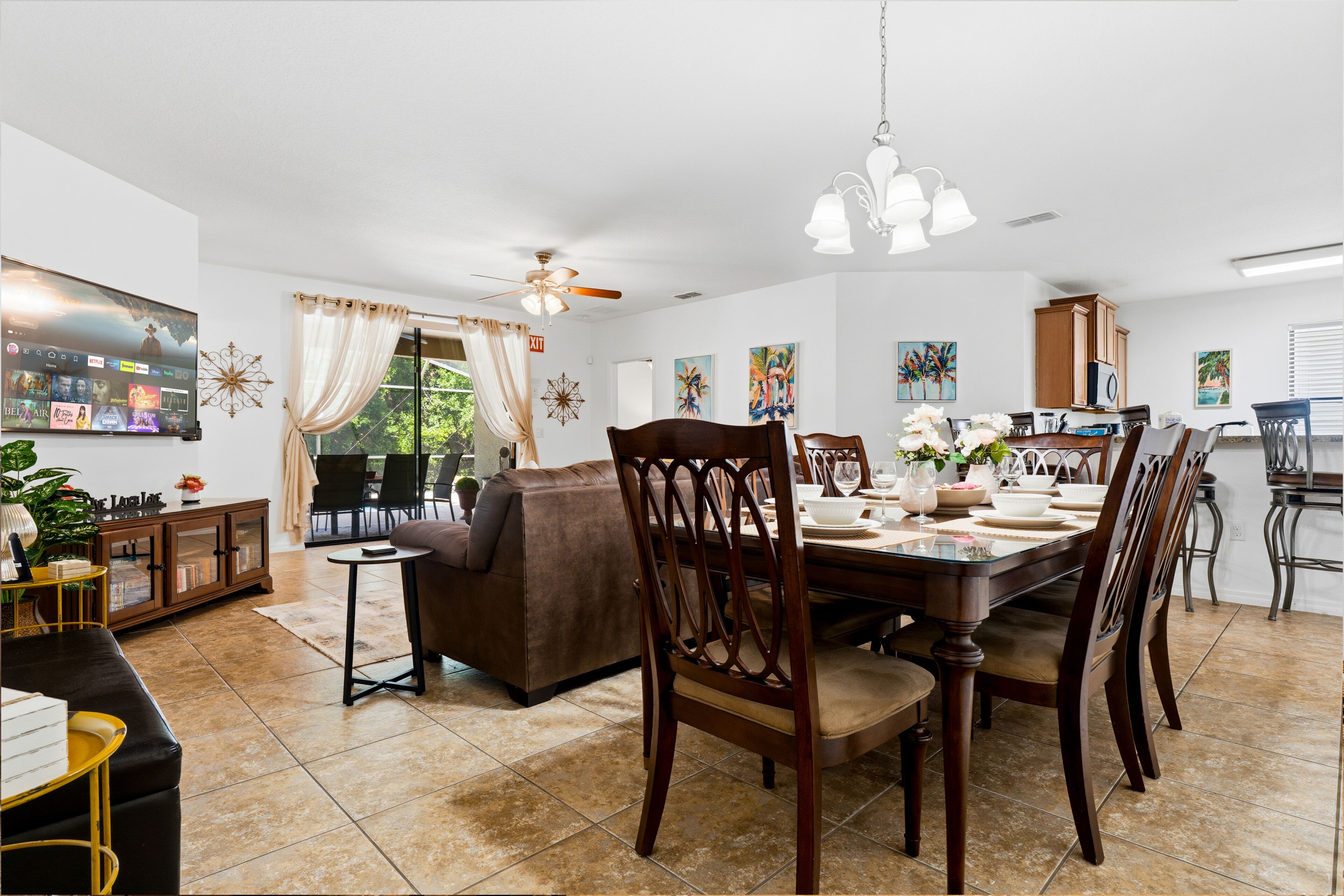 Un comedor con una mesa lista para cuatro personas, rodeada de sillas de madera, un ventilador de techo y una araña.