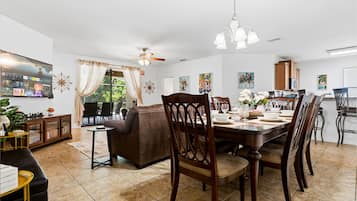 Un comedor con una mesa lista para cuatro personas, rodeada de sillas de madera, un ventilador de techo y una araña.
