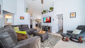 A modern living room with a grey sofa, a grey armchair, and a small ottoman. There is a ceiling fan, a flat-screen TV mounted on the wall, and a small table with a plant.