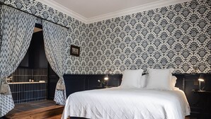 A bedroom with a large bed, dark headboard, patterned wallpaper, and a wooden floor. - Deluxe double room (Le Pian-Médoc)