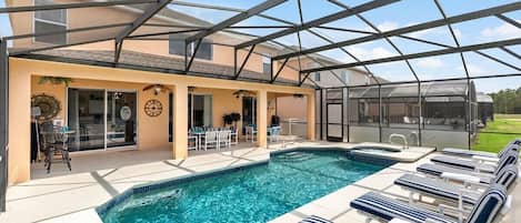 A covered outdoor swimming pool with lounge chairs and a hot tub, surrounded by a house with a patio and a greenhouse.