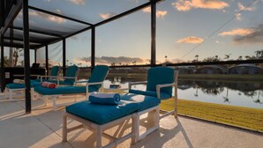 Un espace de détente extérieur, baigné de lumière, avec des fauteuils bleus rembourrés et une vue sur un lac et un quartier résidentiel, le tout protégé par des parois vitrées.