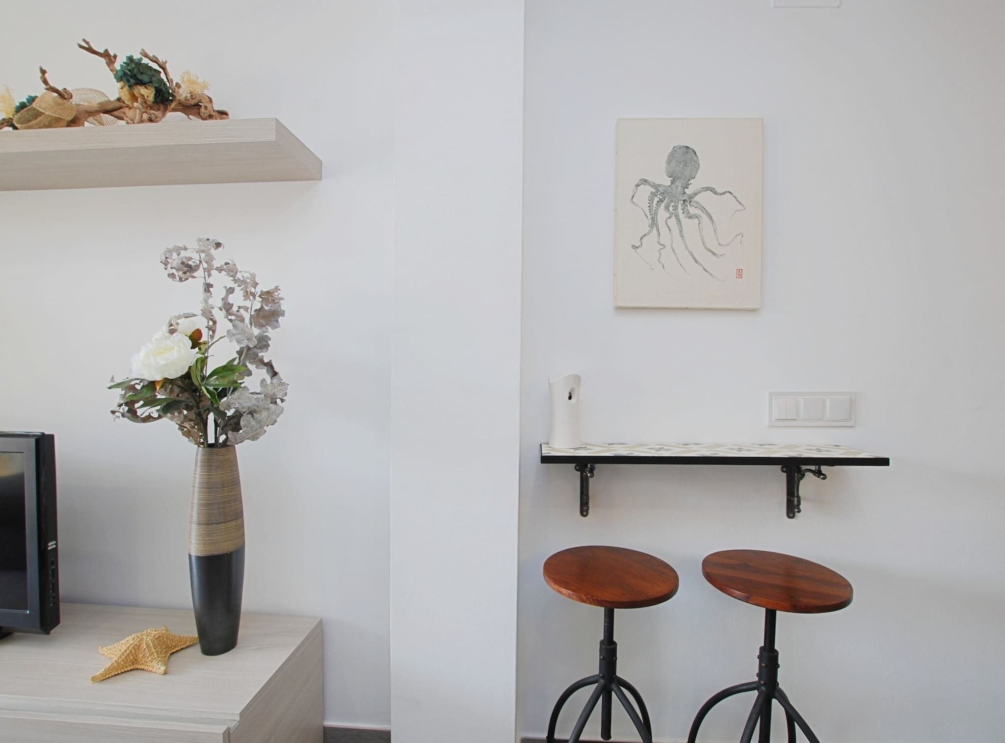 Ein Zimmer mit einem Holztisch, einer Blumenvase, einem Seestern, einem Regal mit einer Zeichnung und zwei Hocker.
