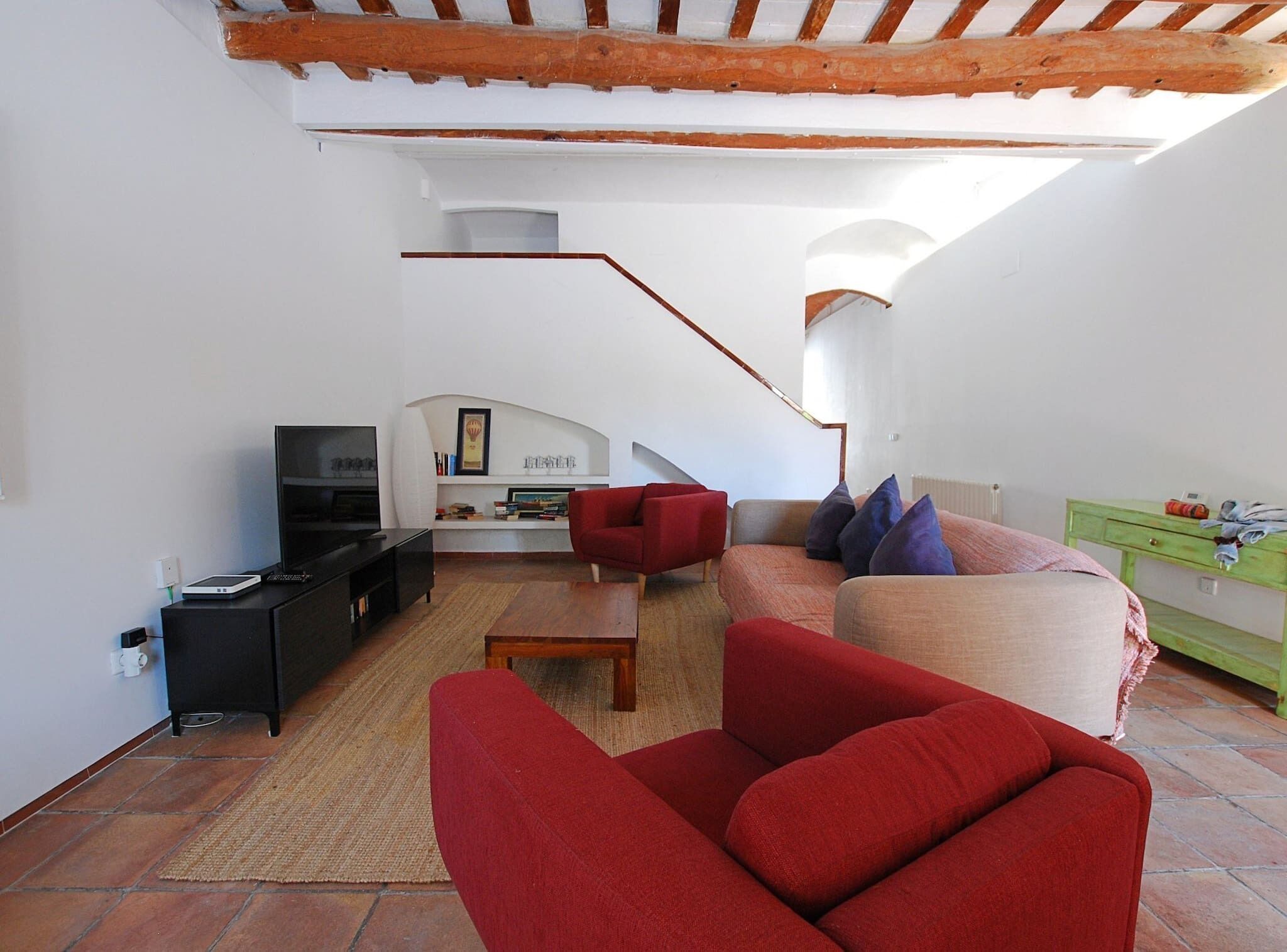 A modern living room with a red sofa, a wooden coffee table, and a TV on a black stand. There is a staircase leading to an upper level.