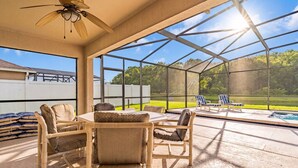 A covered outdoor area with a table and chairs, a ceiling fan, and a view of a swimming pool and greenery. - Lake View Retreat, South-Facing Pool, Spa & Game Room – Quiet Community (Kissimmee)