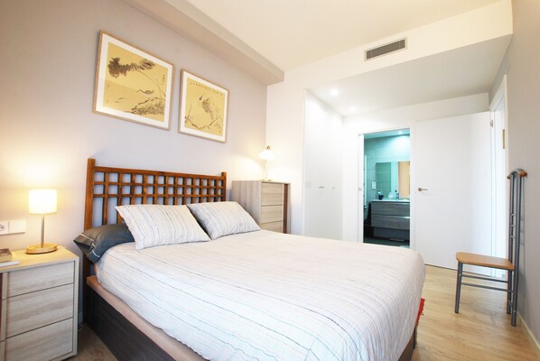 Un dormitorio ordenado con cabecera de madera, mesita de noche con lámpara y una silla.
