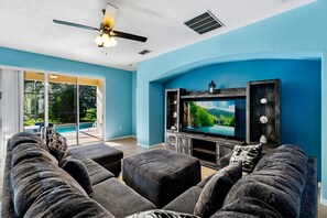 Sala de estar com uma televisão de tela plana grande, sofás cinza escuro e um ventilador de teto.