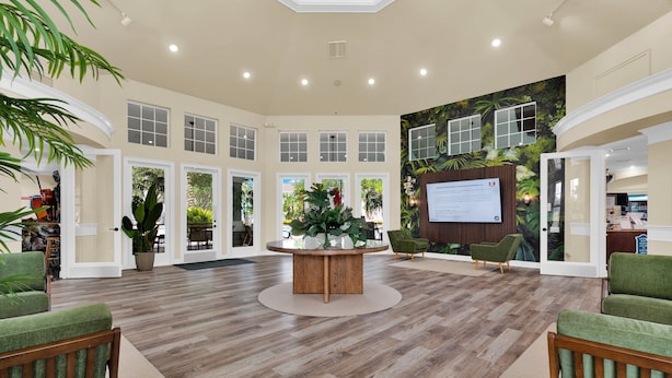 A spacious lobby with a central wooden table, green upholstered chairs, and a tropical-themed wall mural.