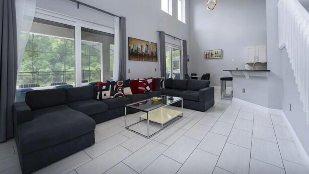 A modern living room with a sectional sofa, a glass coffee table, and large windows offering a view of greenery outside.
