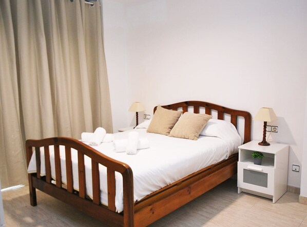 A neatly made bed with white linens and two beige pillows, a wooden headboard, two bedside tables with lamps, and a small plant on one of the tables.
