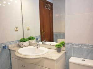 A bathroom with a white sink, a mirror, a wooden door, and two potted plants on the countertop.