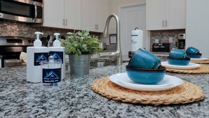 A kitchen with white cabinets, a stainless steel microwave, a sink, and a coffee maker. There are blue ceramic bowls on woven placemats, a potted plant, and a dispenser of white dishwashing soap. - Modern 5BR Oasis w/ Pool, Spa & Resort Perks (Davenport)