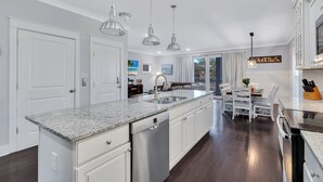Una cocina moderna con armarios blancos, encimeras de granito, electrodomésticos de acero inoxidable, iluminación colgante y una zona de comedor con mesa y sillas.