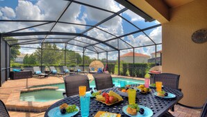 A covered outdoor dining area with a pool, lounge chairs, and a table set with food and drinks.