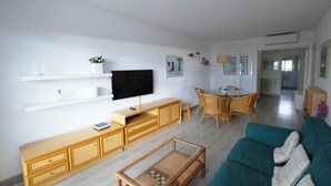 A modern living room with a flat-screen TV, wooden furniture, a teal sofa, and a dining area with wicker chairs.