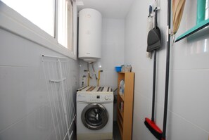A compact laundry room with a front-loading washing machine, a water heater, and a broom on a wall-mounted rack. - Narciso views - Community Pool (Vilassar de Mar)