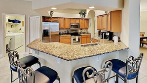 A modern kitchen with granite countertops, stainless steel appliances, and wooden cabinets. There are two bar stools and a built-in island with a sink. - Spacious 4BR Villa with Private Pool Near Disney (Davenport)