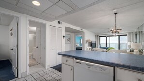A modern kitchen with white cabinetry, a blue countertop, and a dishwasher. There is a hallway leading to a bathroom and a dining area with a view. - Wonderful Ocean Views from this 2 Bedroom Oceanfront With a Pool (Ocean City)