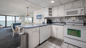 A modern kitchen with white cabinets, a dishwasher, a microwave, and an oven. There is a dining area with a blue table and chairs. - Wonderful Ocean Views from this 2 Bedroom Oceanfront With a Pool (Ocean City)