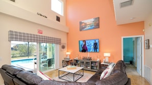 A spacious living area with a large leather sofa, a coffee table, and a television mounted on the wall. There is a pool visible through the glass doors, and an exit sign is visible on the wall. - Disney Villa Free BBQ, Pool & Game Room (Davenport)