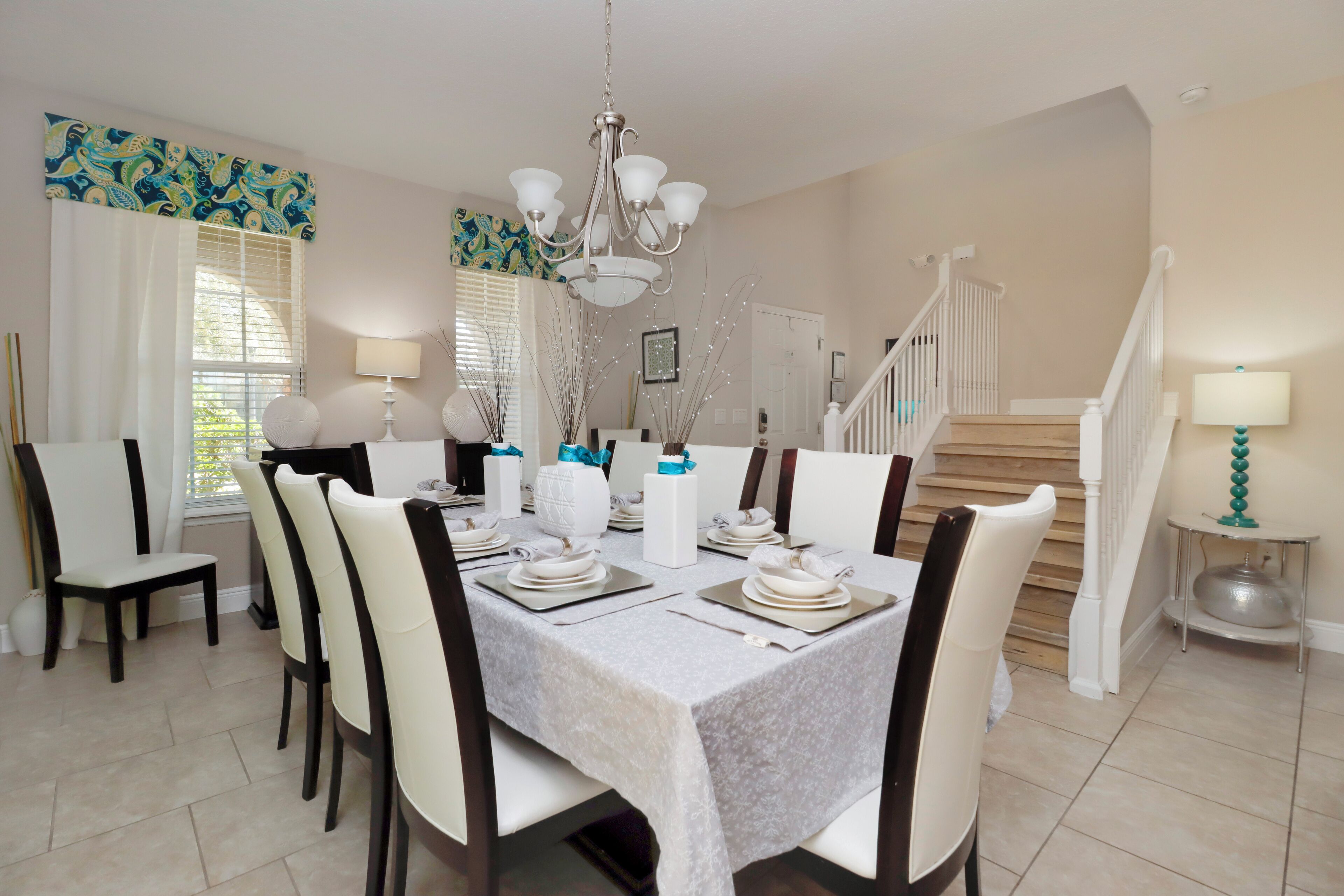 Un coin repas avec une table dressée pour six personnes, des chaises blanches, un lustre et un escalier en arrière-plan.