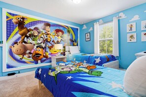 Ein Schlafzimmer mit einer großen Wandmalerei einer Zeichentrickfigur, einem Bett mit blauem Bettbezug und Sternenmuster, einem Nachttisch mit Lampe und einem Fenster mit Vorhängen.