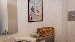 Un baño con una obra de arte enmarcada, una cesta de mimbre y una toalla sobre la encimera.