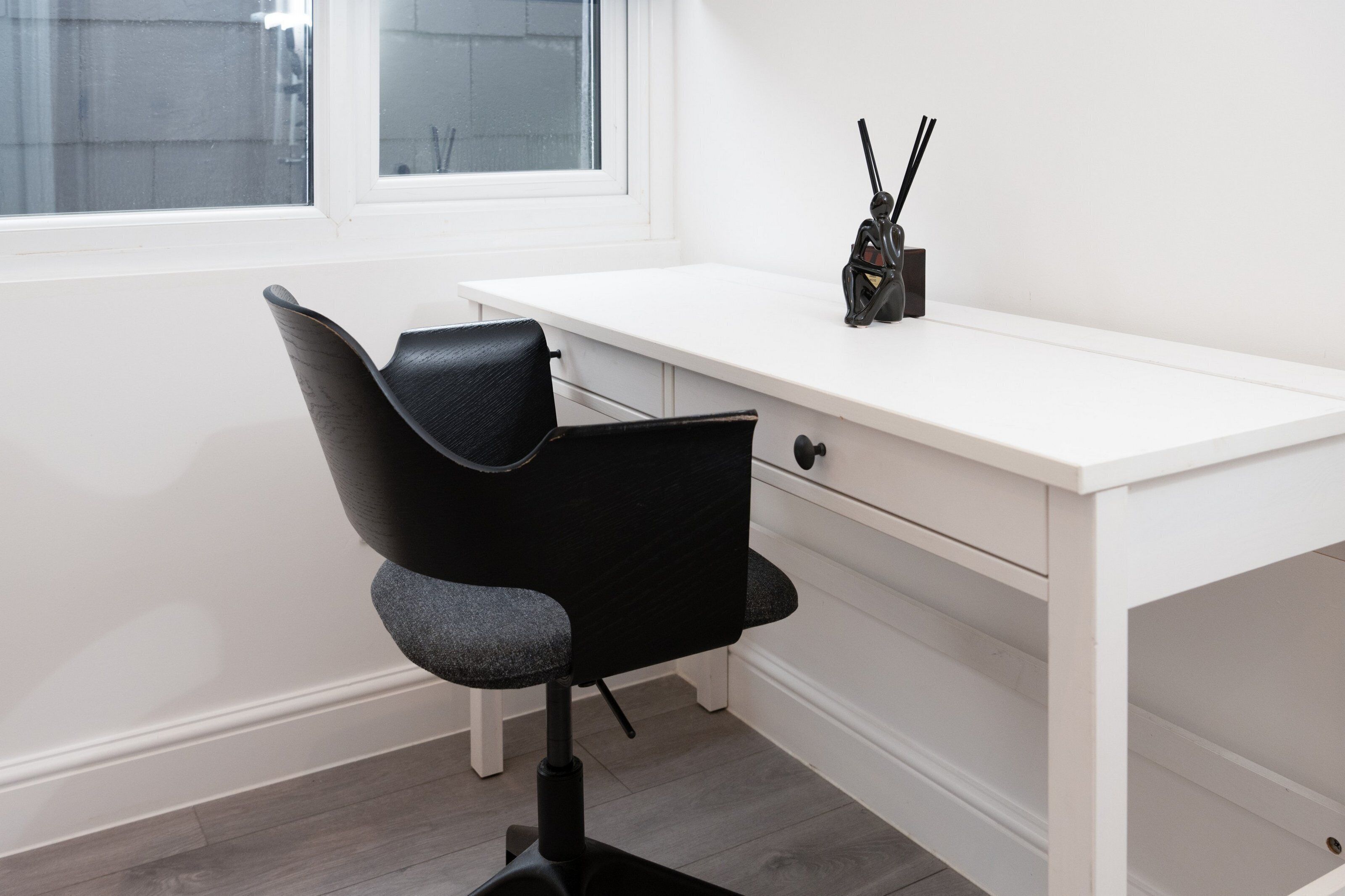 Un bureau blanc avec une chaise noire, une fenêtre et un petit objet noir posé sur le bureau.