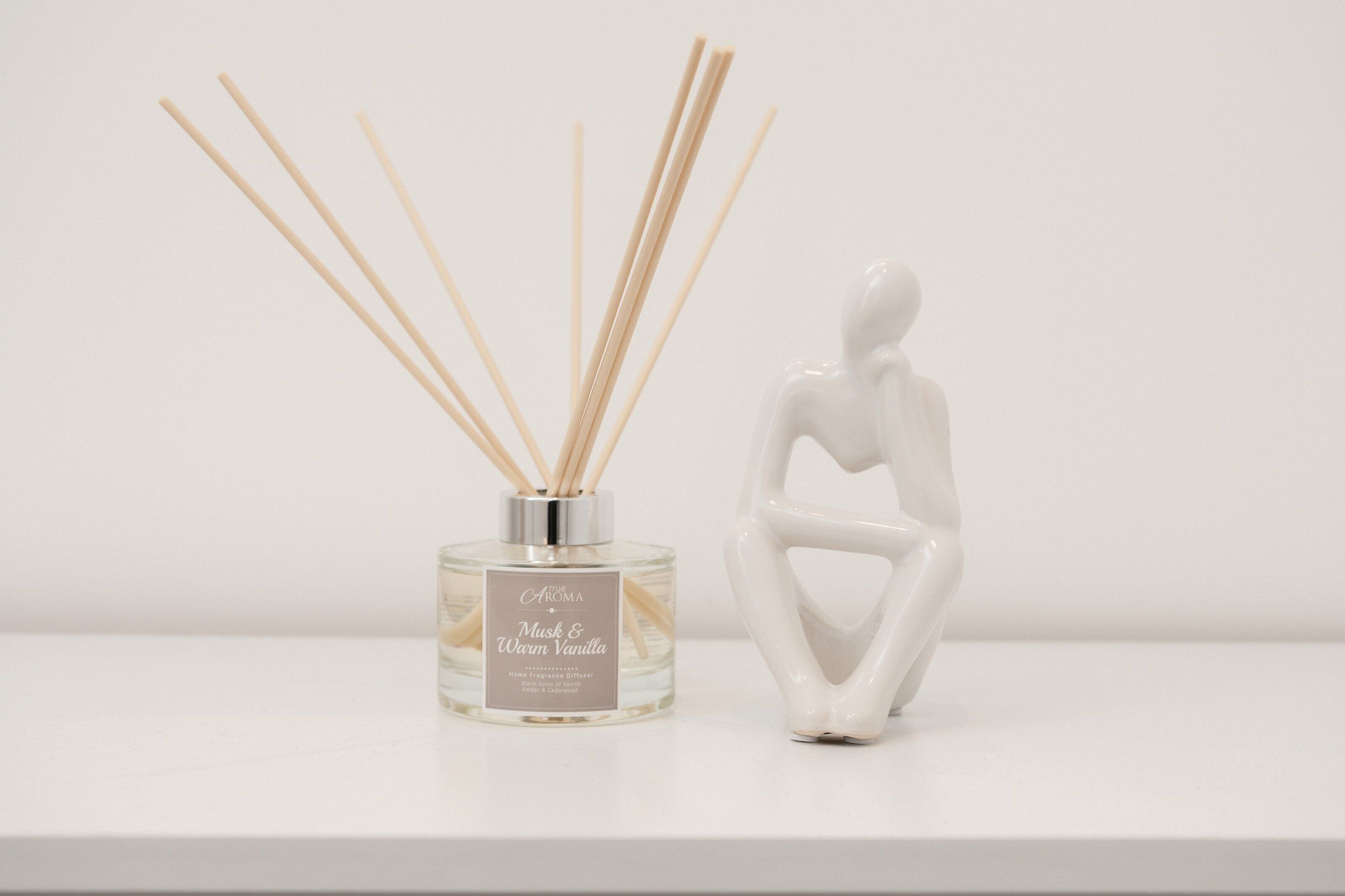 Un difusor de aroma con tiras, 'The Aroma Musk & Warm Vanilla', y una figurita de cerámica blanca sobre una superficie blanca.