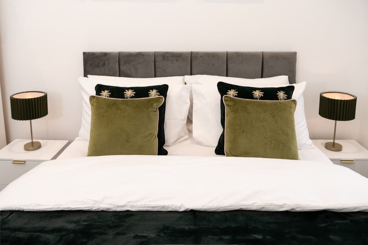 A neatly made bed with white and green pillows, two bedside tables with lamps, and a grey upholstered headboard.
