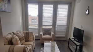 Una sala de estar con sofá, sillón, mesa de centro, televisor y ventanas amplias con persianas.