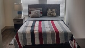 Un dormitorio con cama, dos ventanas con persianas, ventilador de techo, mesita de noche, lámpara y una silla.