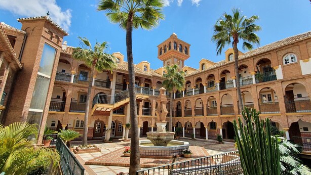 Un patio central con una fuente, rodeado de edificios de ladrillo con balcones y palmeras.