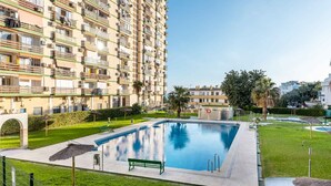 Una piscina con un banco verde, rodeada de un césped cuidado y un edificio residencial alto al fondo.