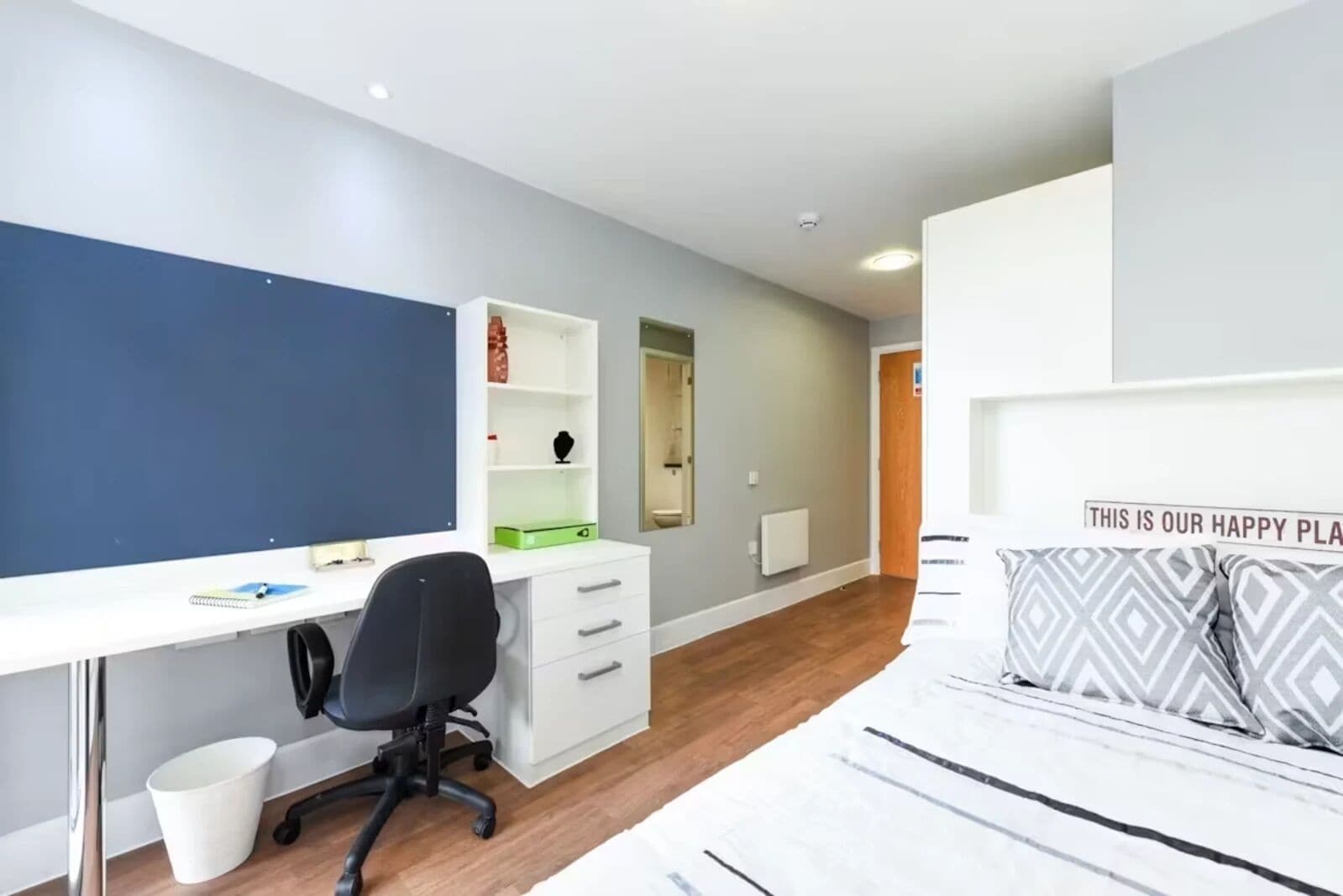 A bedroom with a bed, a desk, a chair, a white dresser, and a shelf with items.