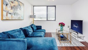 A modern living room with a blue sofa, a flat-screen TV, a painting, and a window with blinds. - Basildon - 2-bed - Sleeps 5 - Near Train Station (Basildon)