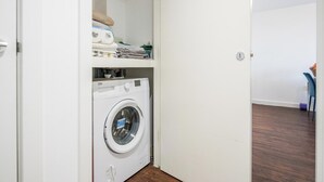 A compact laundry room with a front-loading washing machine, a drying rack, and a shelf with folded clothes and a small appliance. - Basildon - 2-bed - Sleeps 5 - Near Train Station (Basildon)