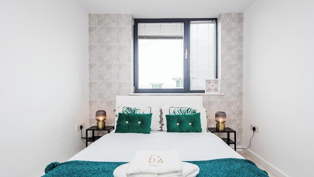 A bedroom with a bed, green pillows, a white towel, and two bedside lamps. - Basildon - 2-bed - Sleeps 5 - Near Train Station (Basildon)