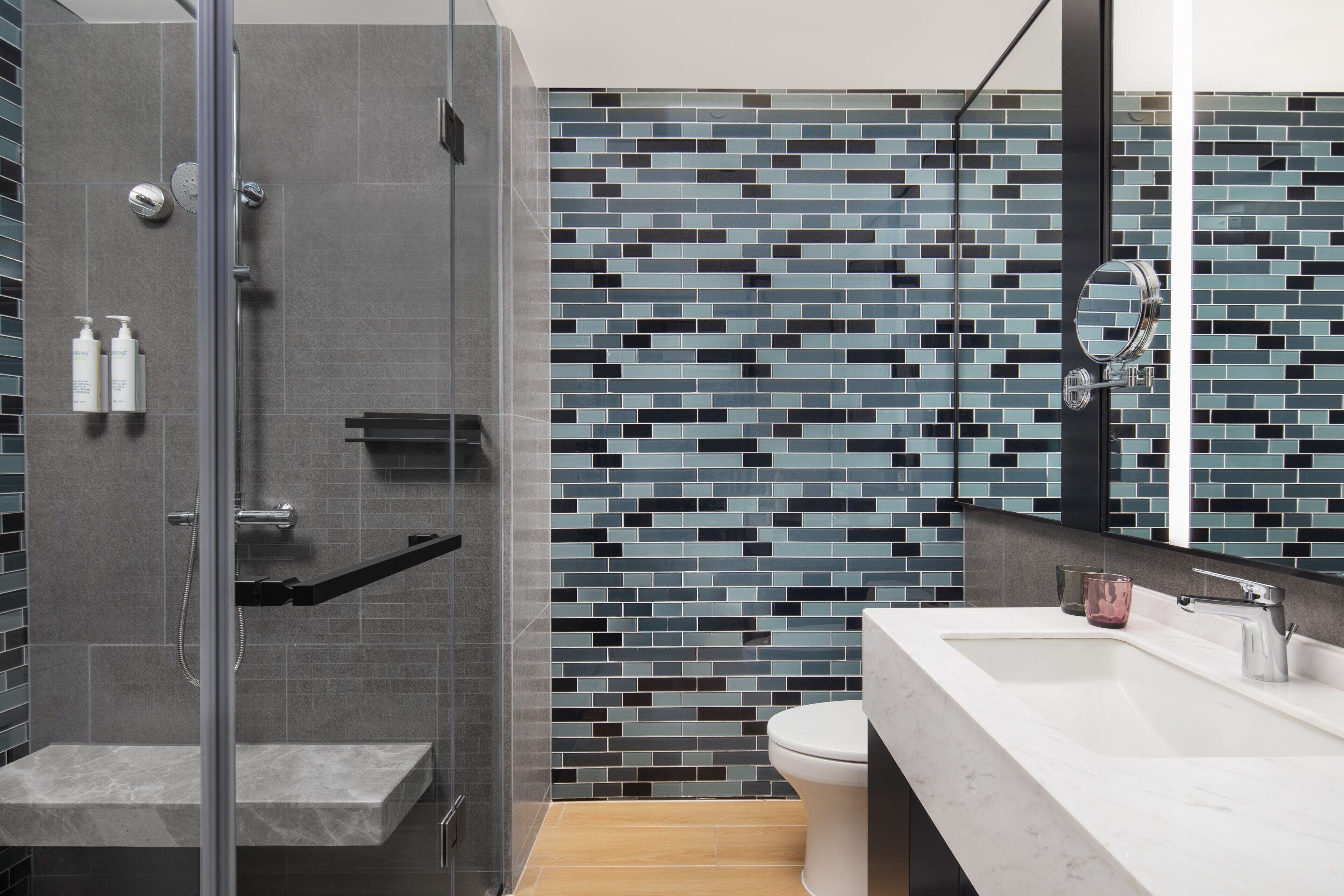 Un baño moderno con ducha envuelta en vidrio, pared con diseño de mosaico y lavamanos blanco con espejo encima.