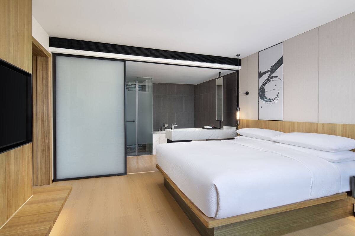 Habitación de hotel moderna con una cama grande, televisión, puerta corredera de vidrio al baño y diseño minimalista.