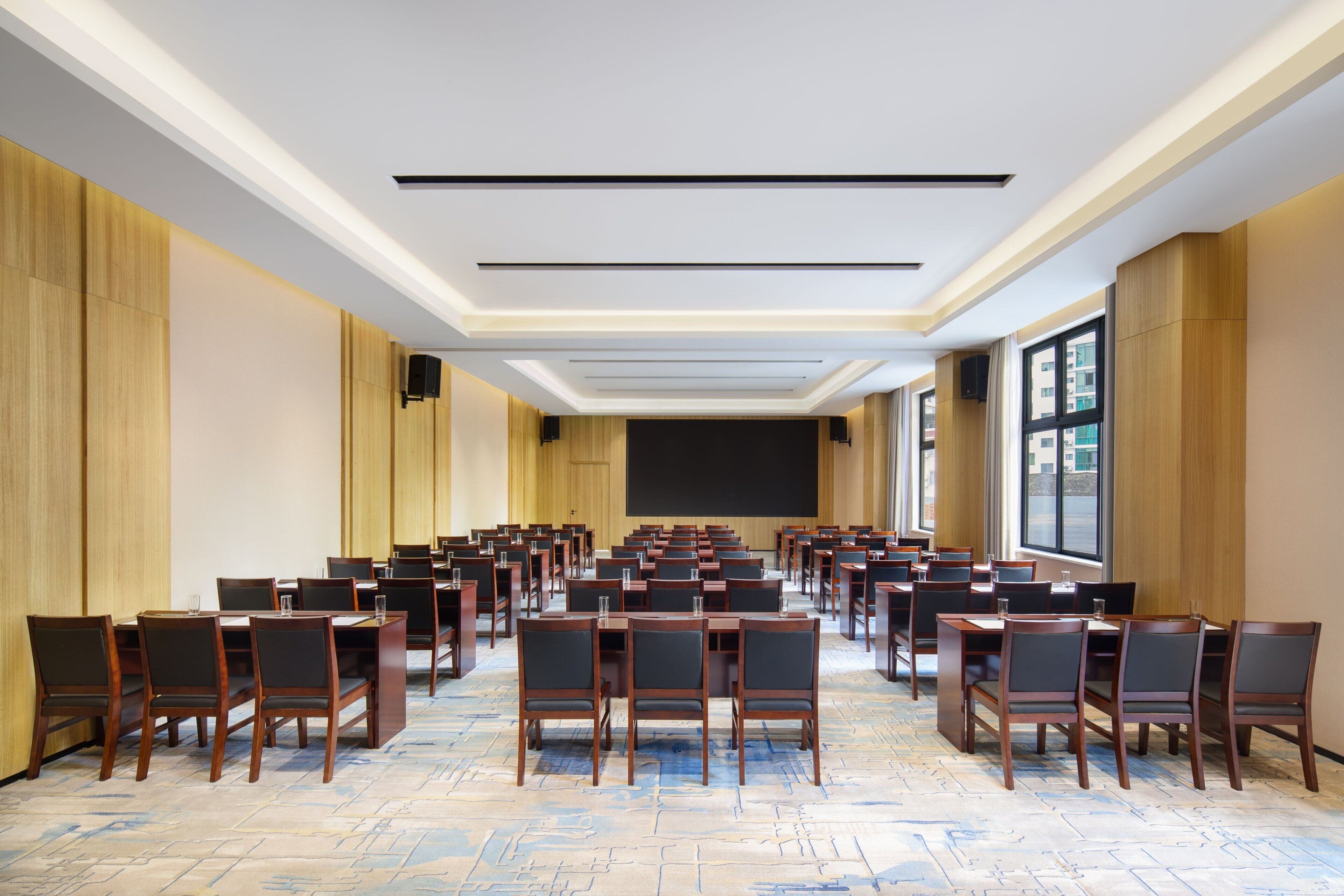Une salle de conférence avec des rangées de tables et de chaises, un grand écran à l’avant et une vue sur les bâtiments à travers de grandes fenêtres.