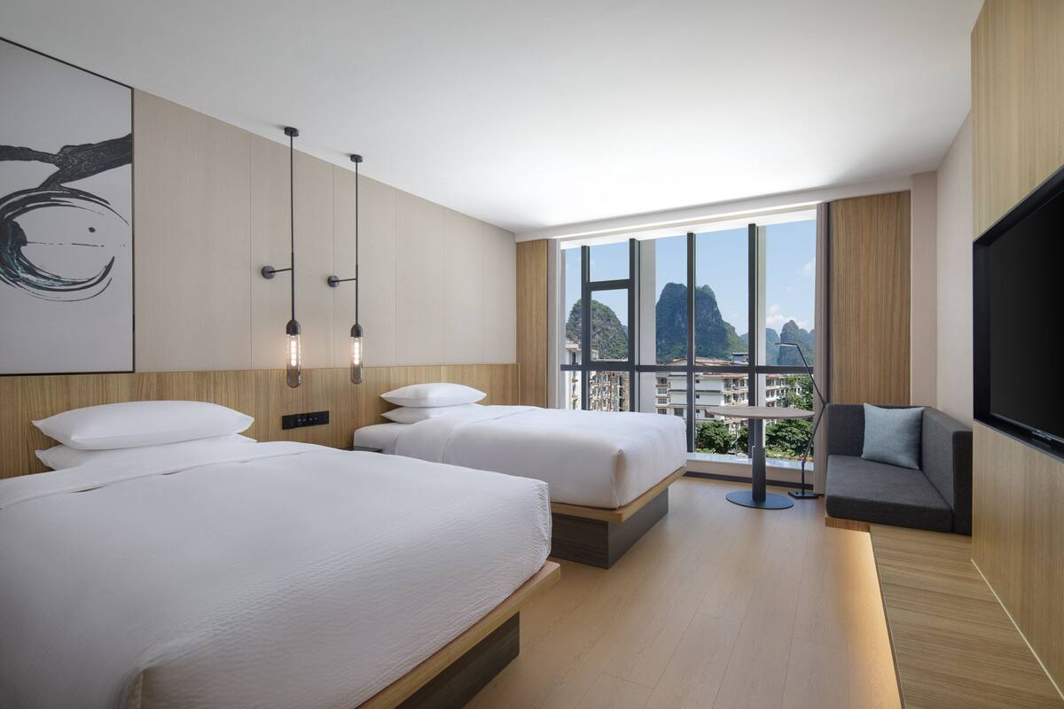 Una habitación de hotel moderna con dos camas, un ventanal grande con vistas a la ciudad y un televisor de pantalla plana.