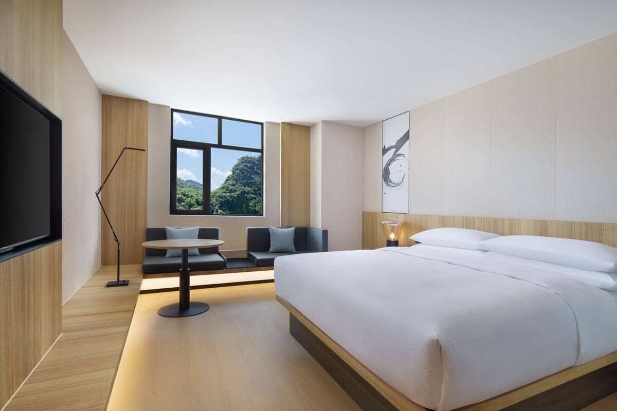 Habitación de hotel moderna con una cama grande, una zona de estar con mesa, televisión y ventana con vistas a zonas verdes.