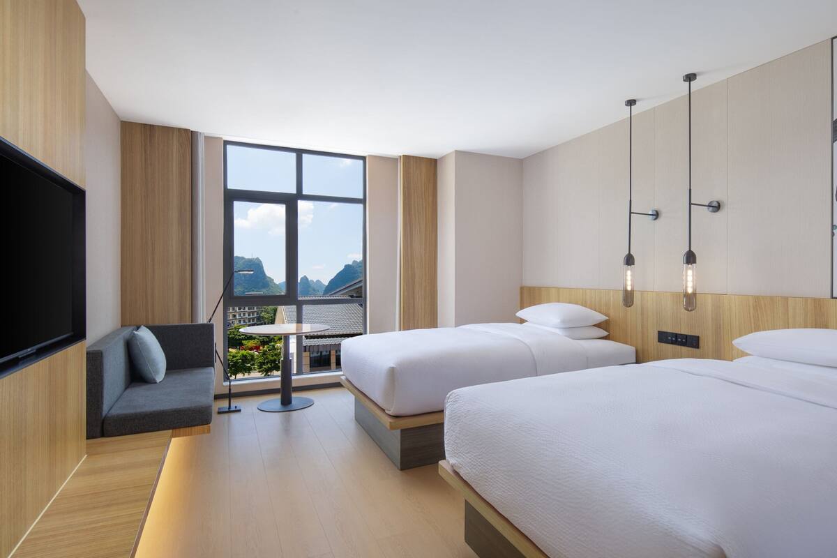 Habitación de hotel moderna con dos camas, televisor, sofá y un amplio ventanal con vistas a la montaña.