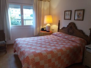 Ein ordentlich bezogenes Bett mit einem gemusterten Überwurf, einer Nachttischlampe, einem Nachttisch und einem Fenster mit Vorhängen.