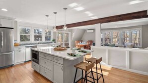 A modern kitchen with a large island, wooden bar stools, and pendant lighting. - Updated A-Frame w/ Game Room, Short Drive to Beach! (Barnstable)